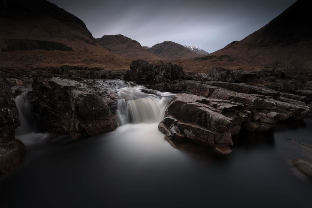 Colour long-exposure winter landscape in Glen Etive, Scotland, with dark peaty water flowing almost stout-like through blocky reddish-pink rocks into a black foreground pool, beneath bare brown hillsides, snow-dusted distant tops and a pale fading sky.