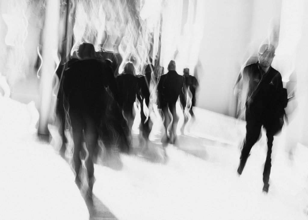 High-key black-and-white street photograph with dark figures walking through a bright white space, surrounded by curling light trails, with one distinct figure on the right.